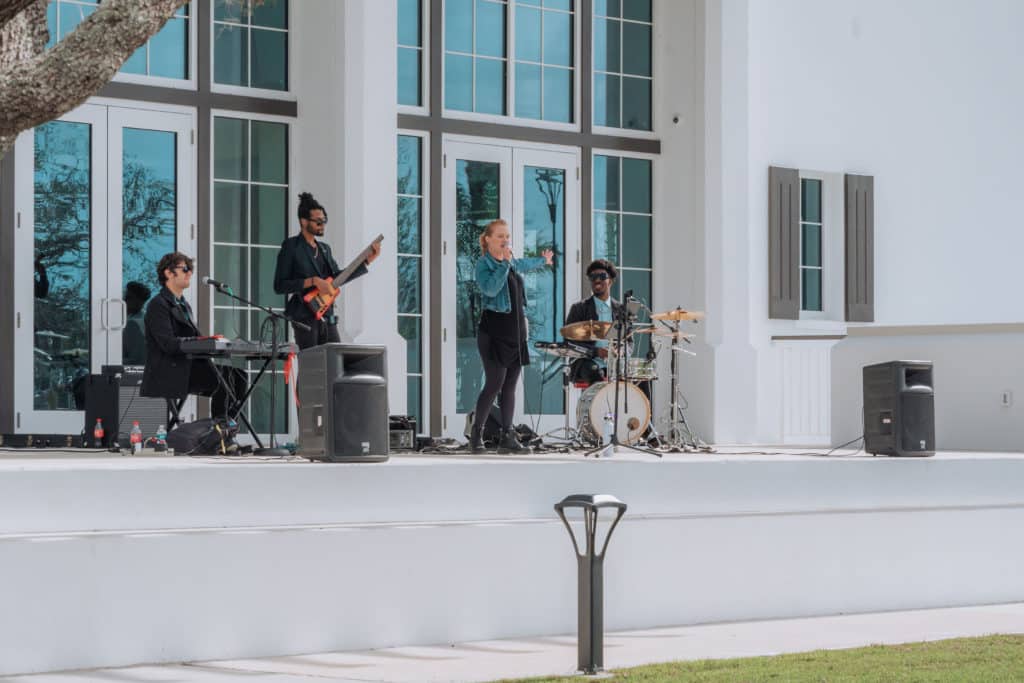 Band members with instruments playing on a white outdoor stage in front of large windows from Solis Hall in Downtown Wellen