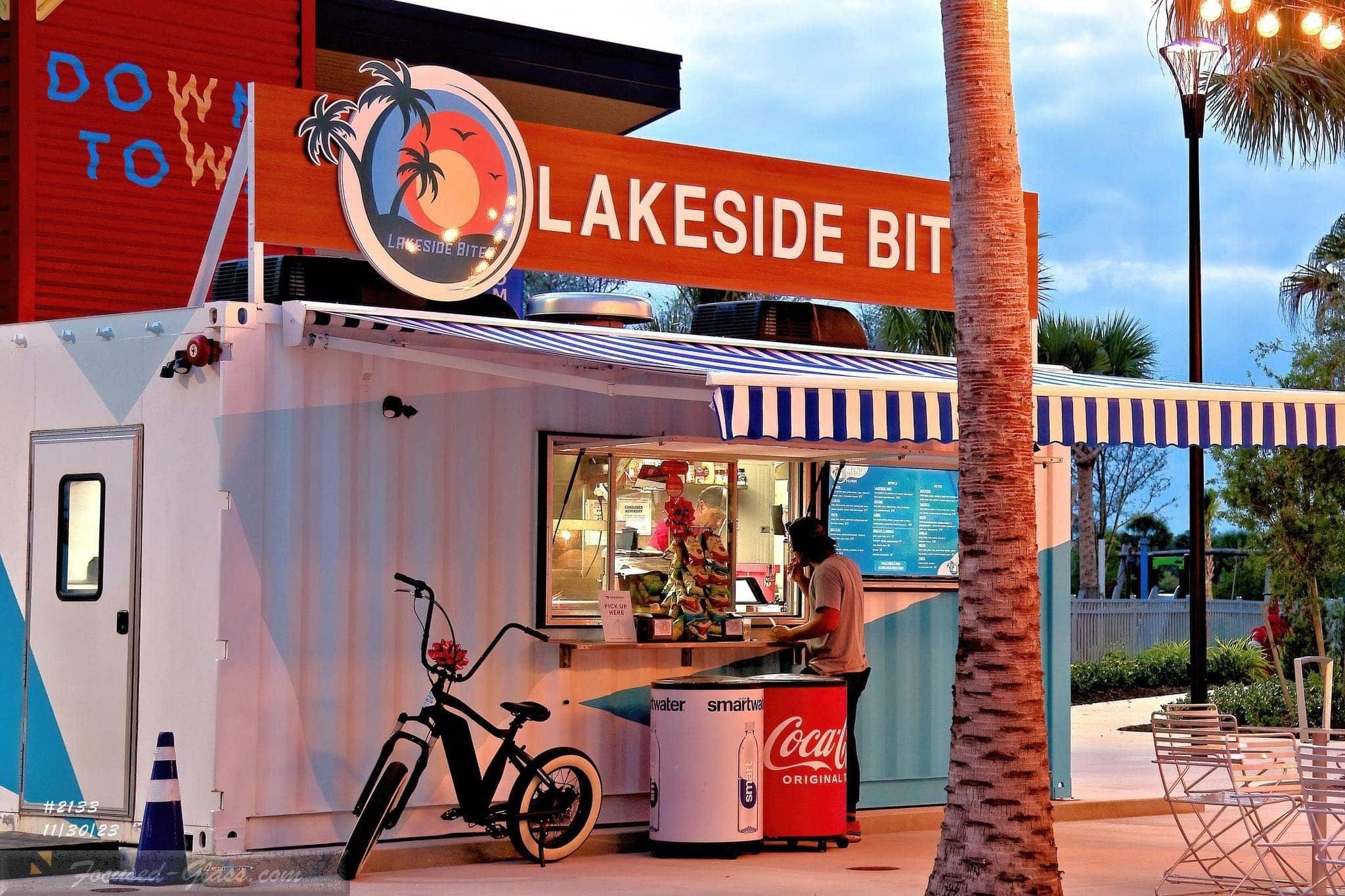 A stand alone, shack called the Lakeside Bites with a bright red roof, pink and blue patterns painted on it and two retro coca-cola branded coolers next to a kids bike out front