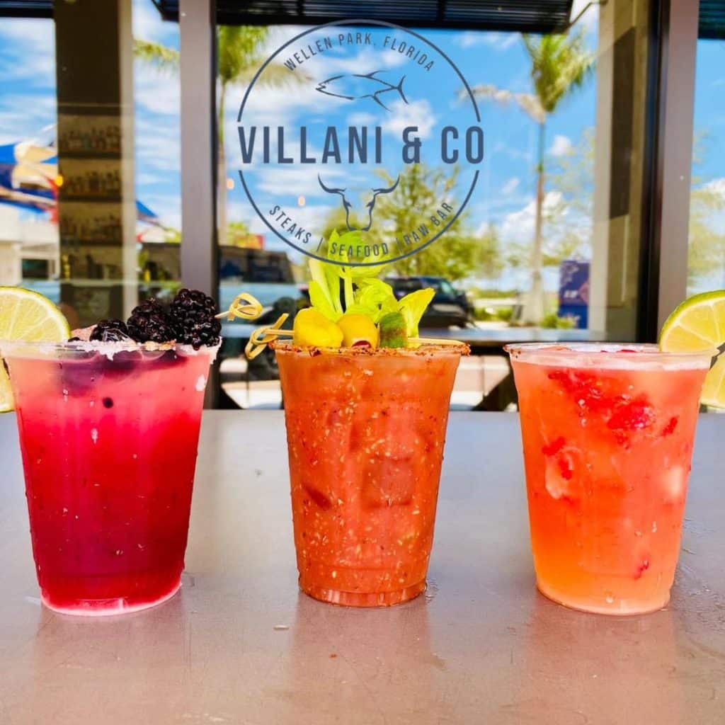 Three red and orange tropical drinks with garnishes sitting on a table. There is a window decal behind that says "Villani & Co" 