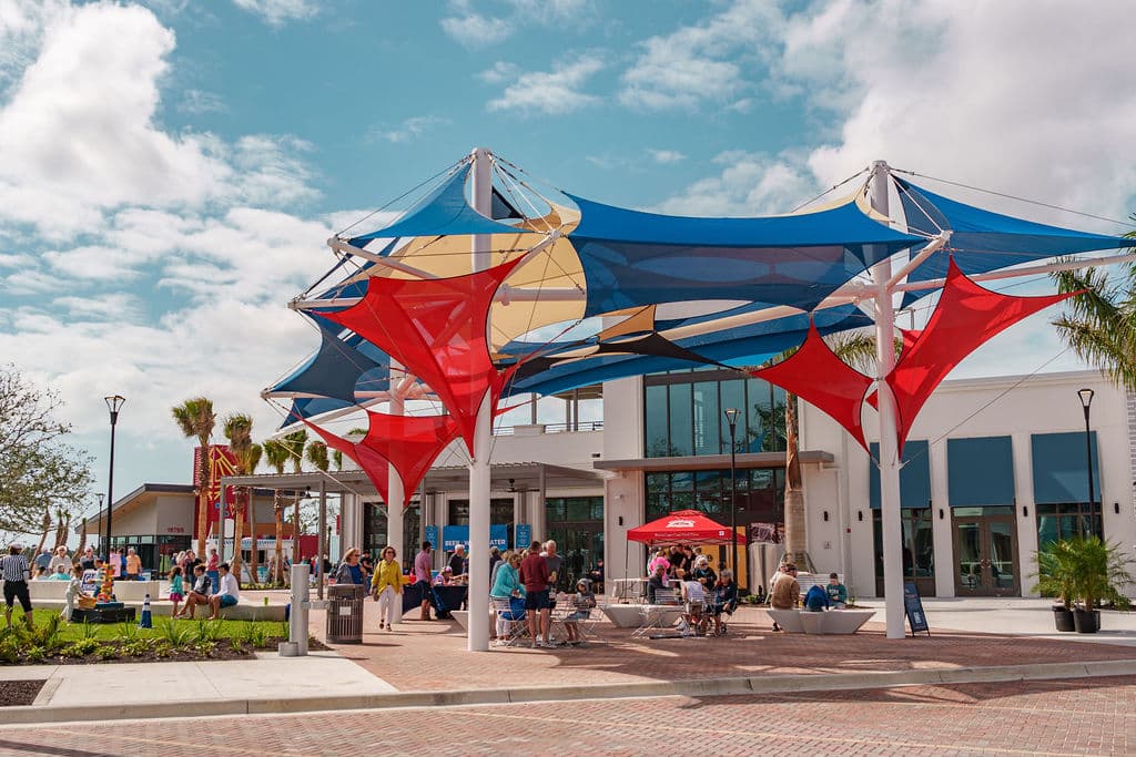 Outdoor plaza with colorful red and blue shade sails, modern architecture, and people gathered under sunny skies.