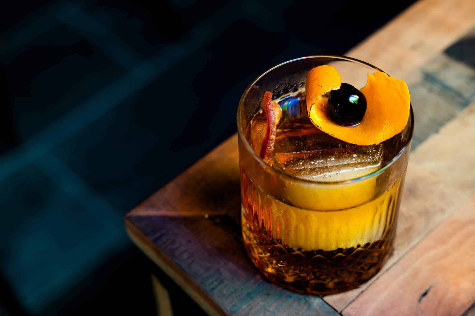 A glass of an old fashion with orange peels and a black cherry on top