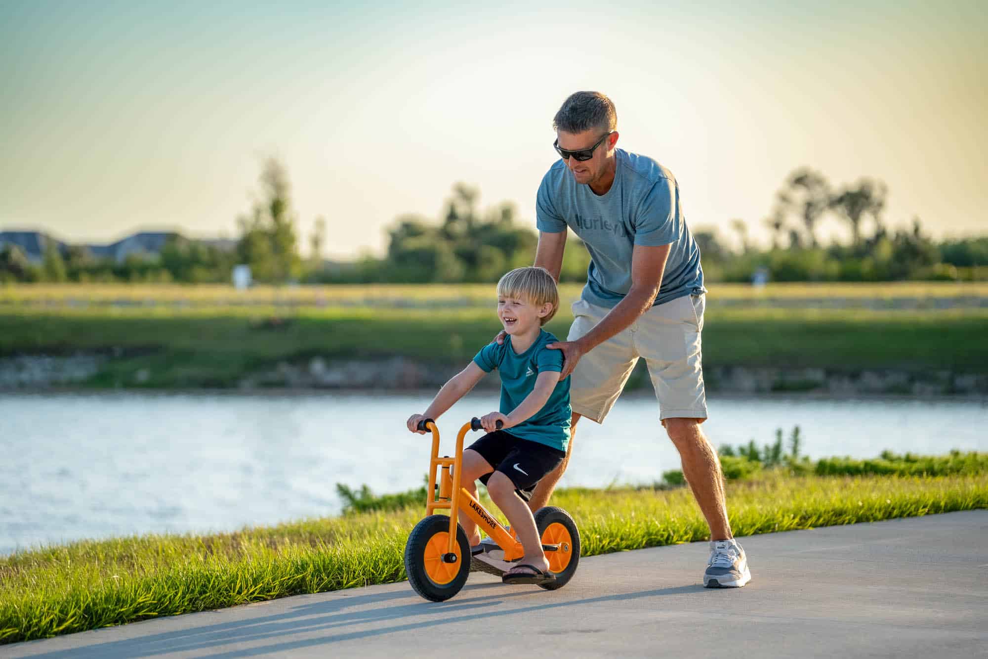 A man pushing a little boy on a small yellow bike on the promenade by the water