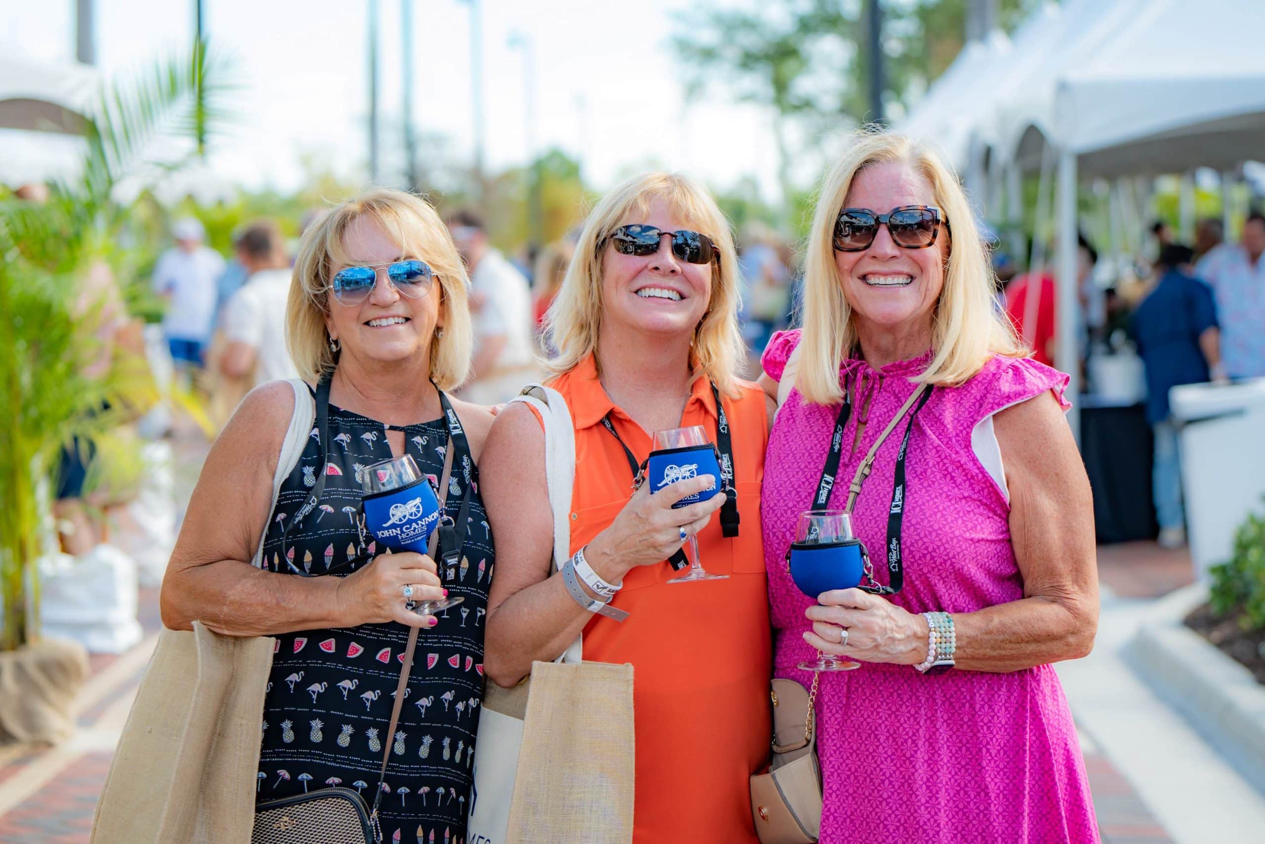 Three women smiling for a picture at an outdoor event, holding covered wine glasses