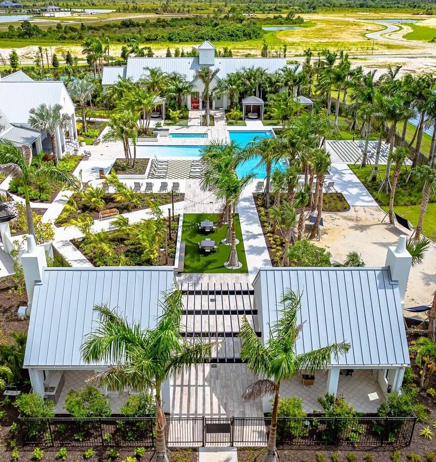 An aerial view of the Everly at Wellen pool area with a covered lounge featuring a kitchen, dining tables, a sand pit with hamocks, all leading up to a pool in the center of several lounging chairs, palm trees and other amenity buildings