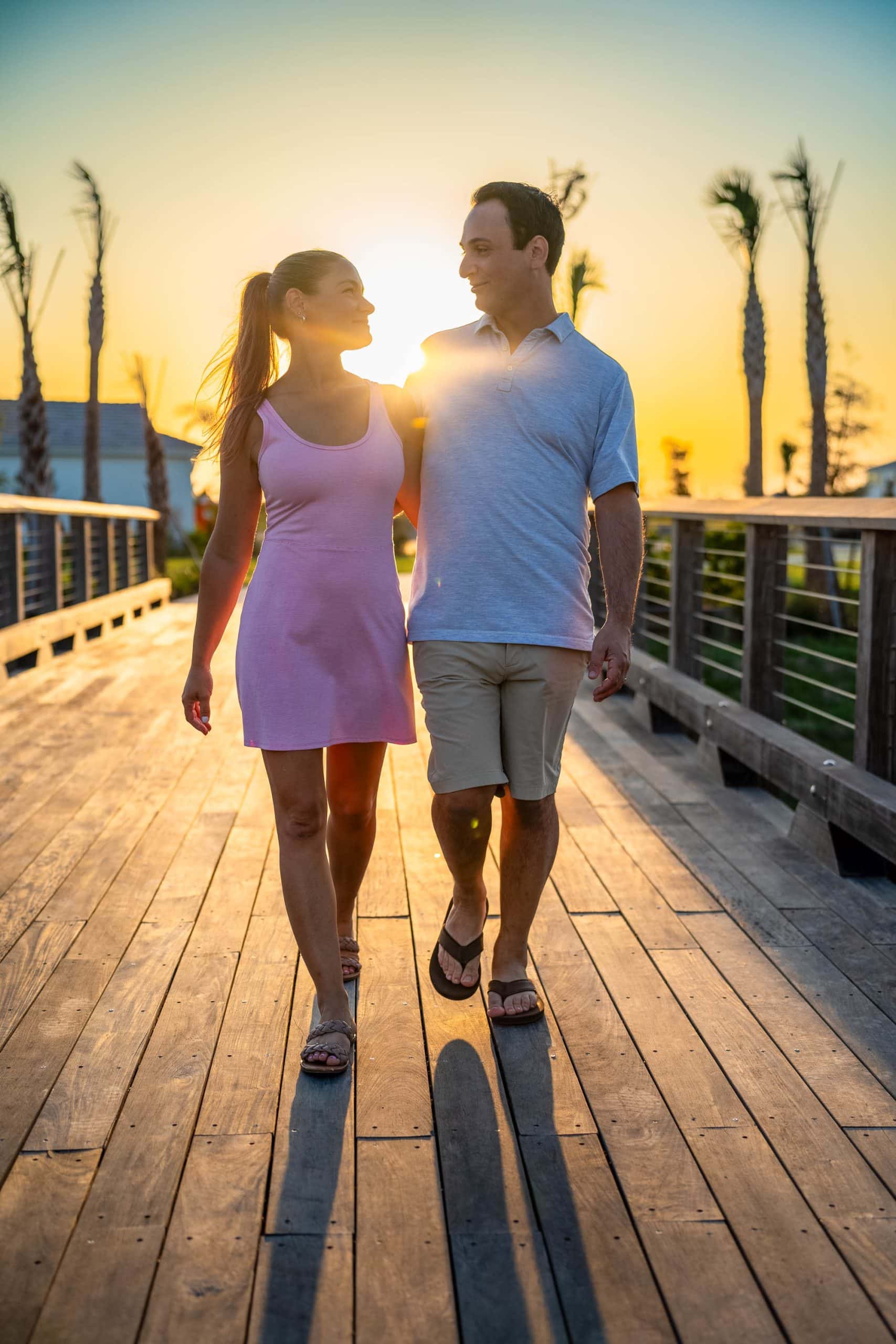 A couple walking in each others arms, crossing a wooden bridge with the sun setting behind them