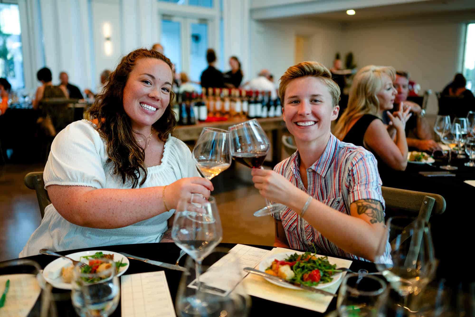 Guests raising glasses in a toast during a wine-tasting event, seated around a table filled with glassware inside a bright, privant event venue.