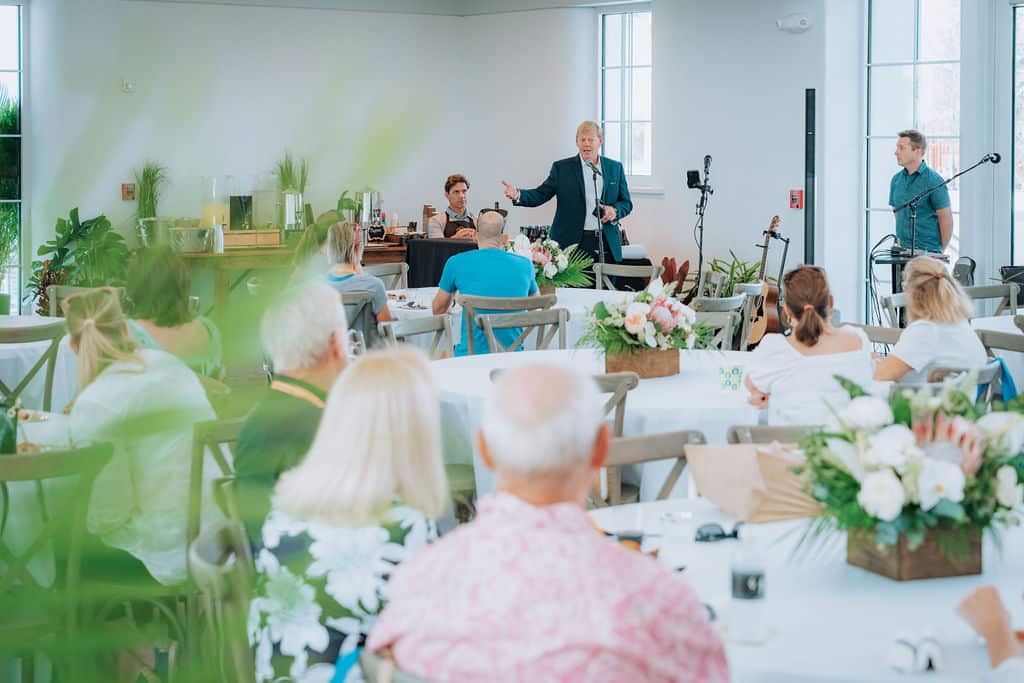 Speaker presenting to an audience seated at round tables with floral centerpieces during a daytime event inside a bright, modern corporate event venue.