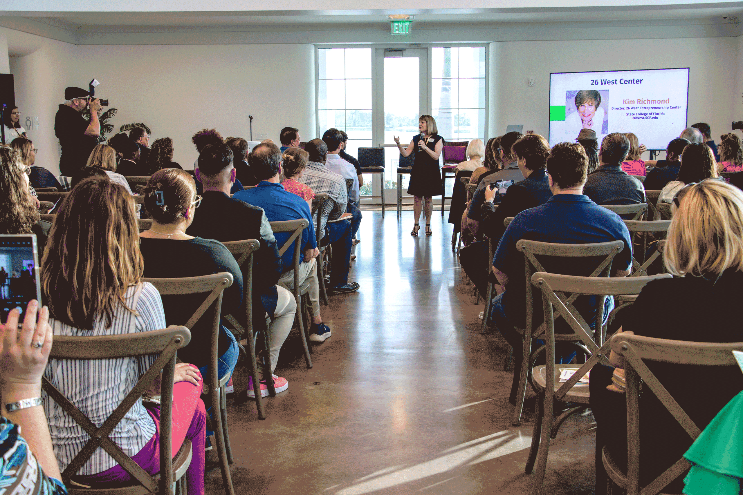 Speaker presenting to a full audience during a business seminar, with attendees seated in rows and a large screen displaying event information inside a bright, modern venue.