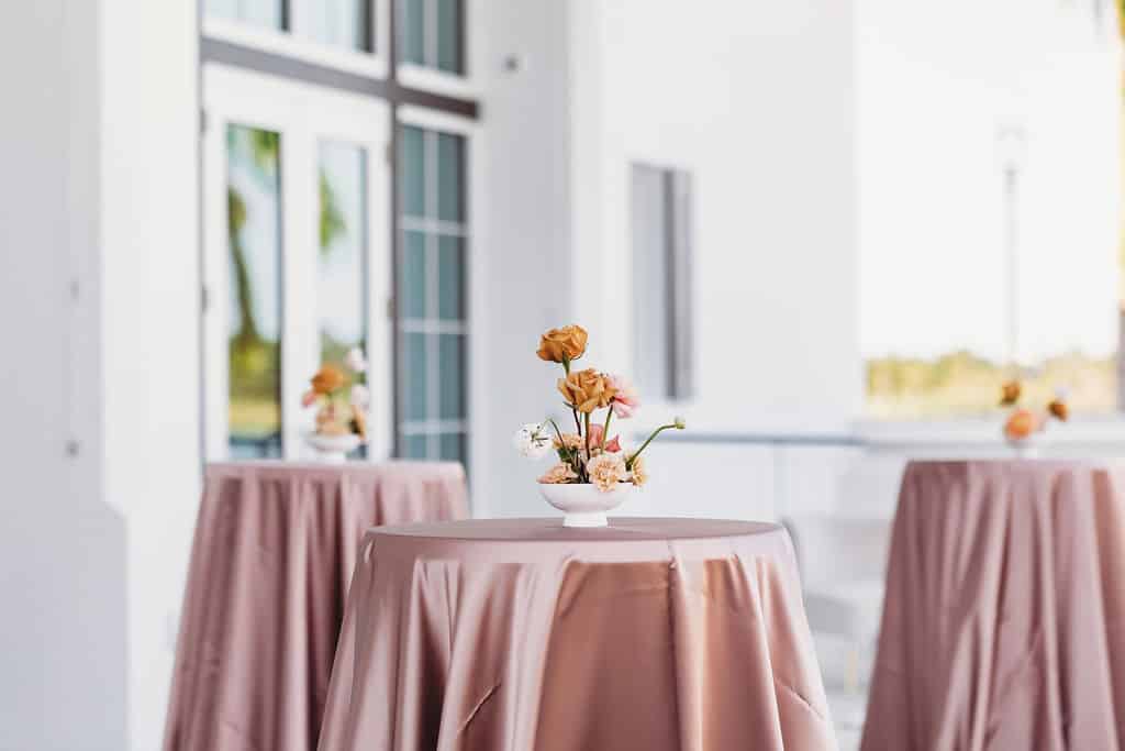 Cocktail-hour tables draped in blush linens with minimalist floral centerpieces arranged on an outdoor patio at a modern wedding private event venue venue.