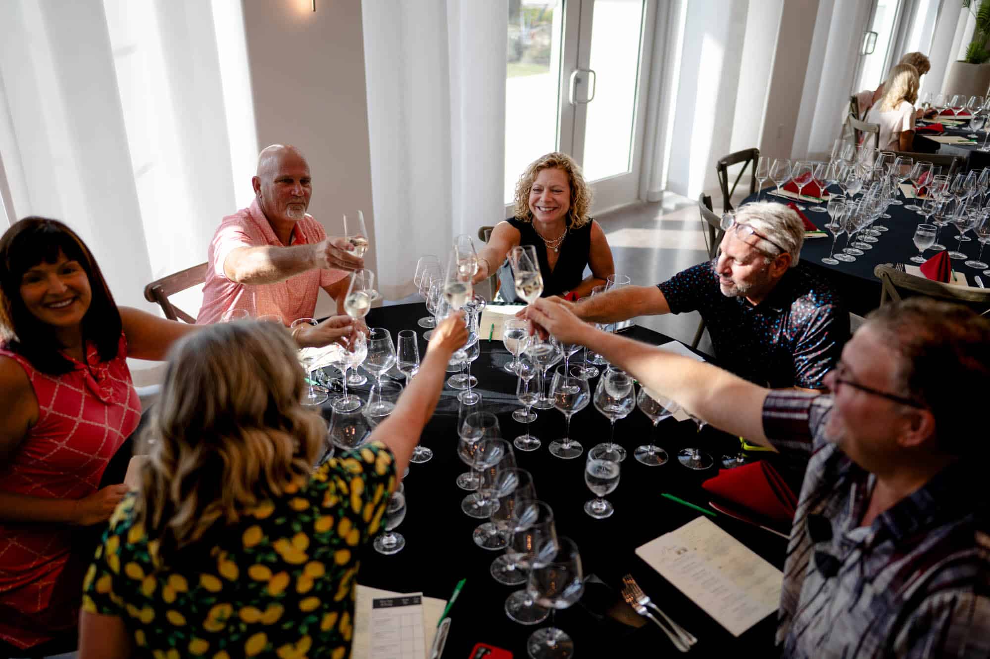 Two guests smiling and clinking wine glasses during a tasting event, seated at a table with plates of food and surrounded by other attendees in a corporate event venue.