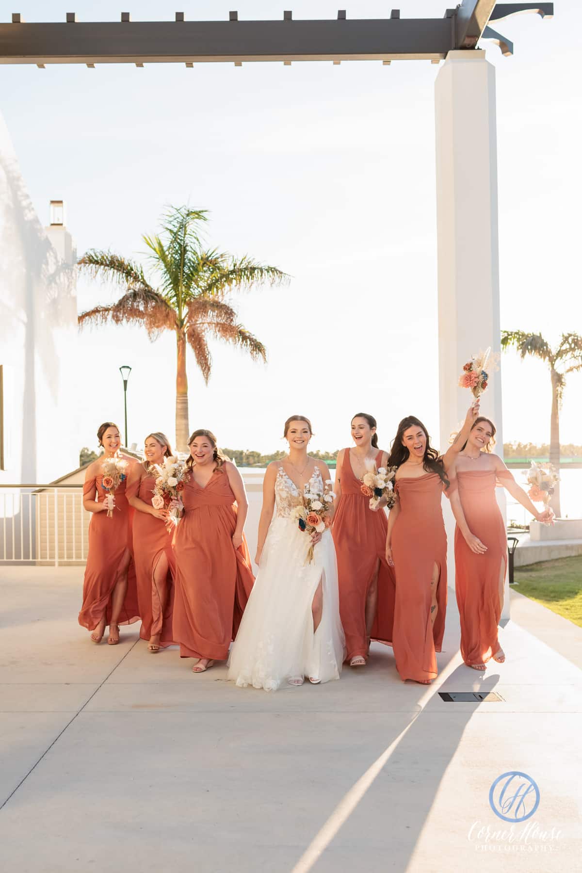 Bride walking with her bridesmaids in matching terracotta dresses, holding floral bouquets during a sunny outdoor wedding near palm trees.