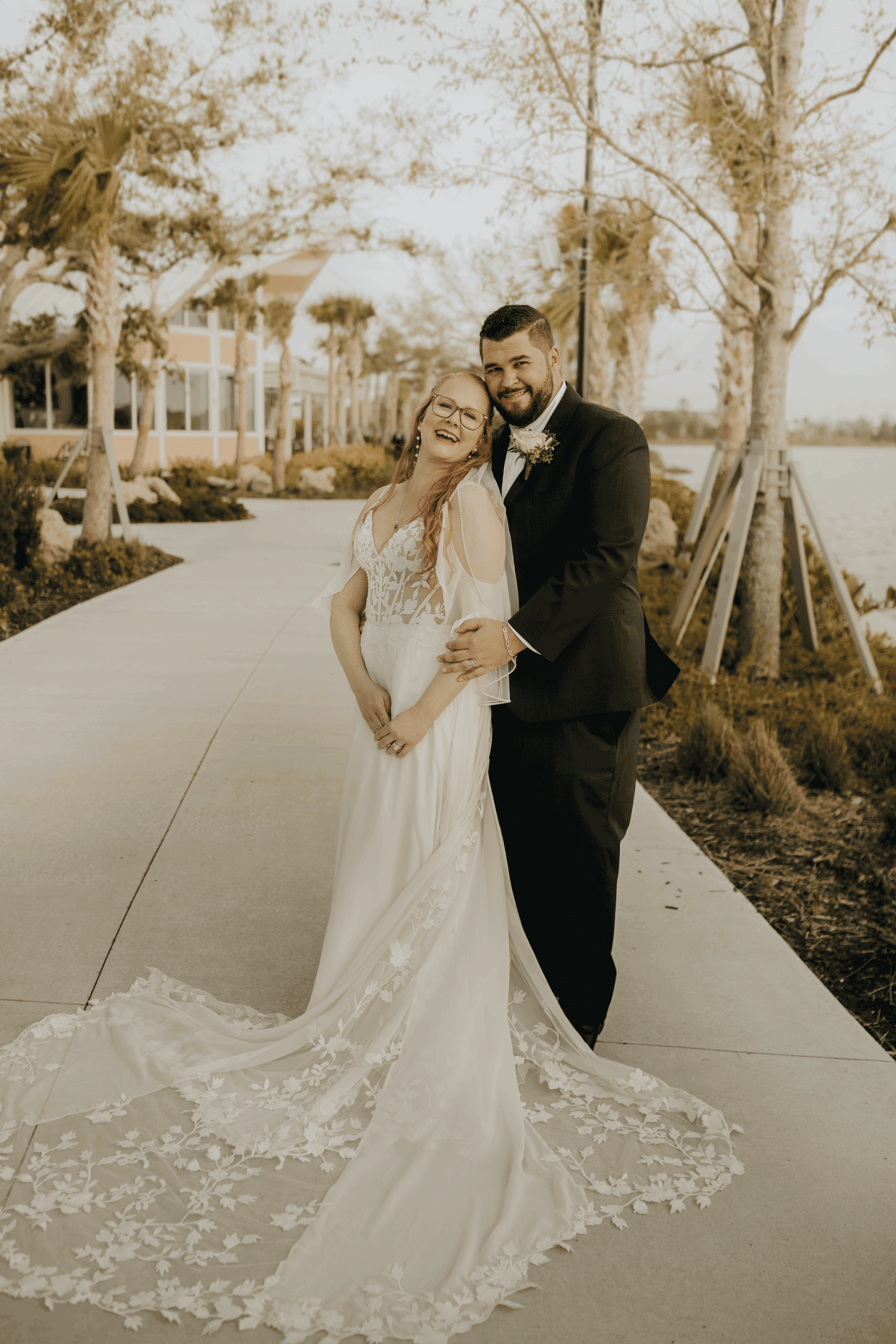 Bride and groom laughing together during golden hour on a lakeside walkway, the bride wearing a lace wedding gown with a long floral train.