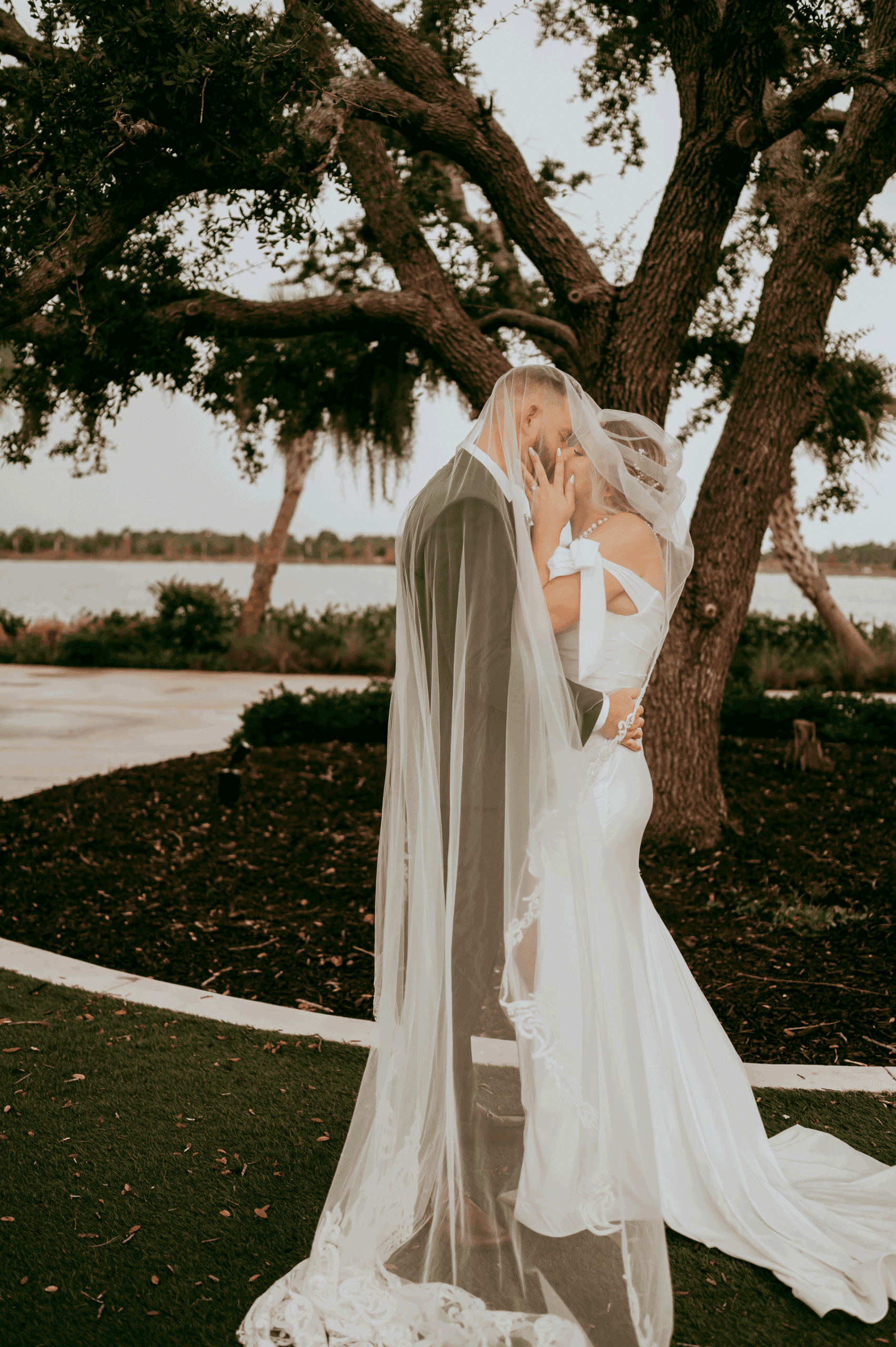 Bride and groom sharing an intimate moment under the veil during their outdoor lakeside wedding, standing beneath a large oak tree