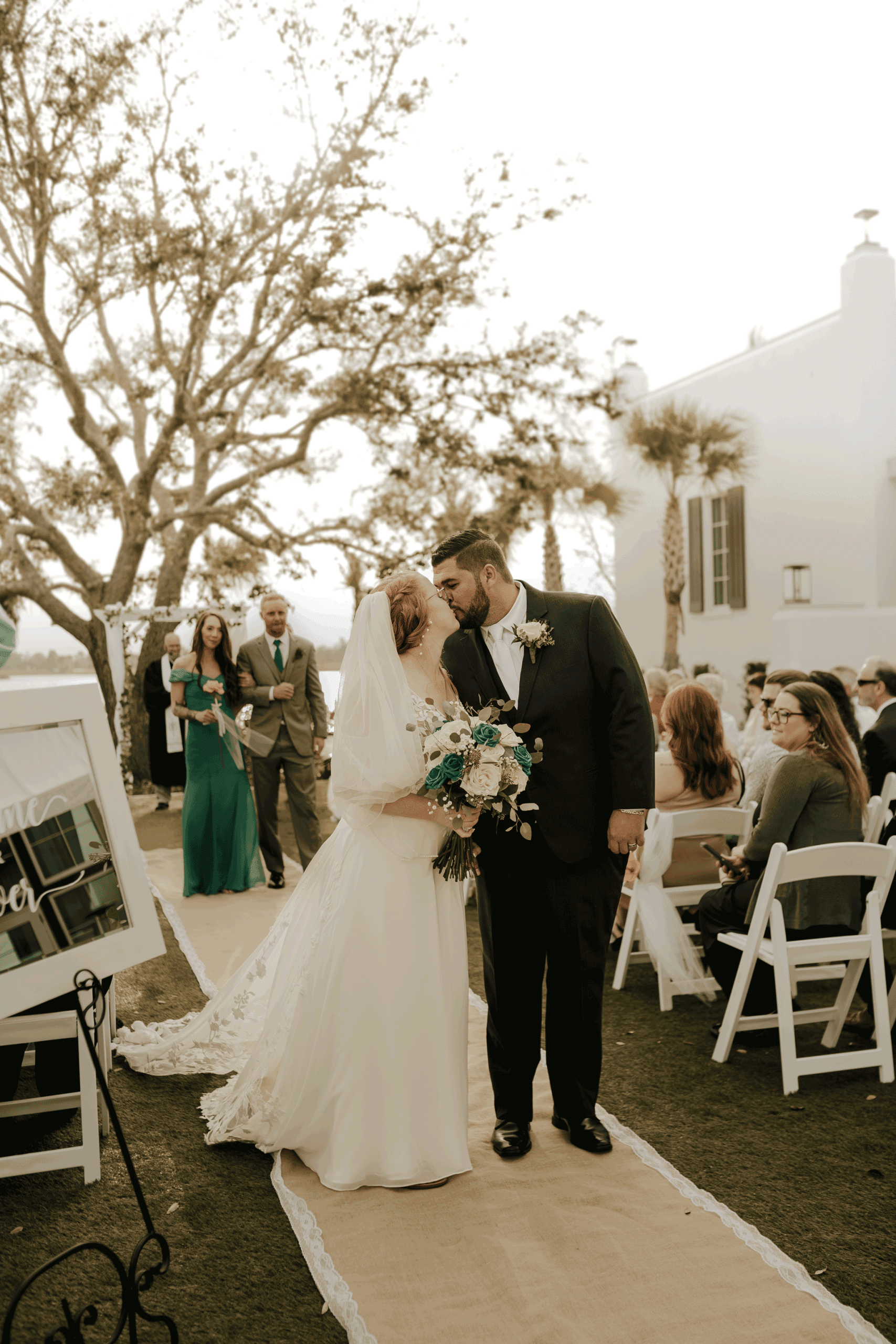 Bride and groom sharing a kiss while walking up the aisle after their outdoor wedding ceremony, surrounded by seated guests and framed by palm trees and a lakeside backdrop.