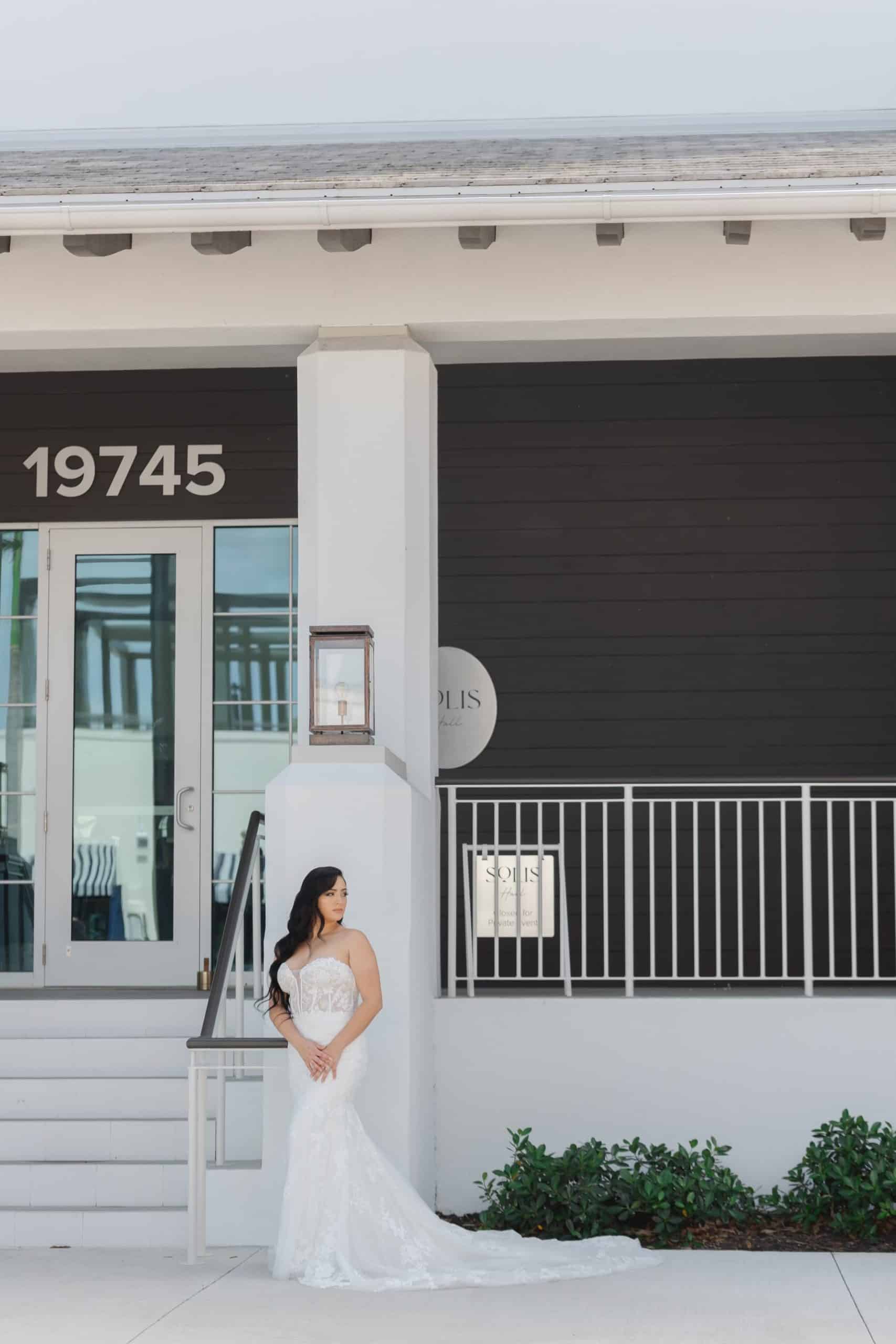 Bride in a strapless lace mermaid gown posing against the modern exterior of a black-and-white wedding venue building with address 19745.