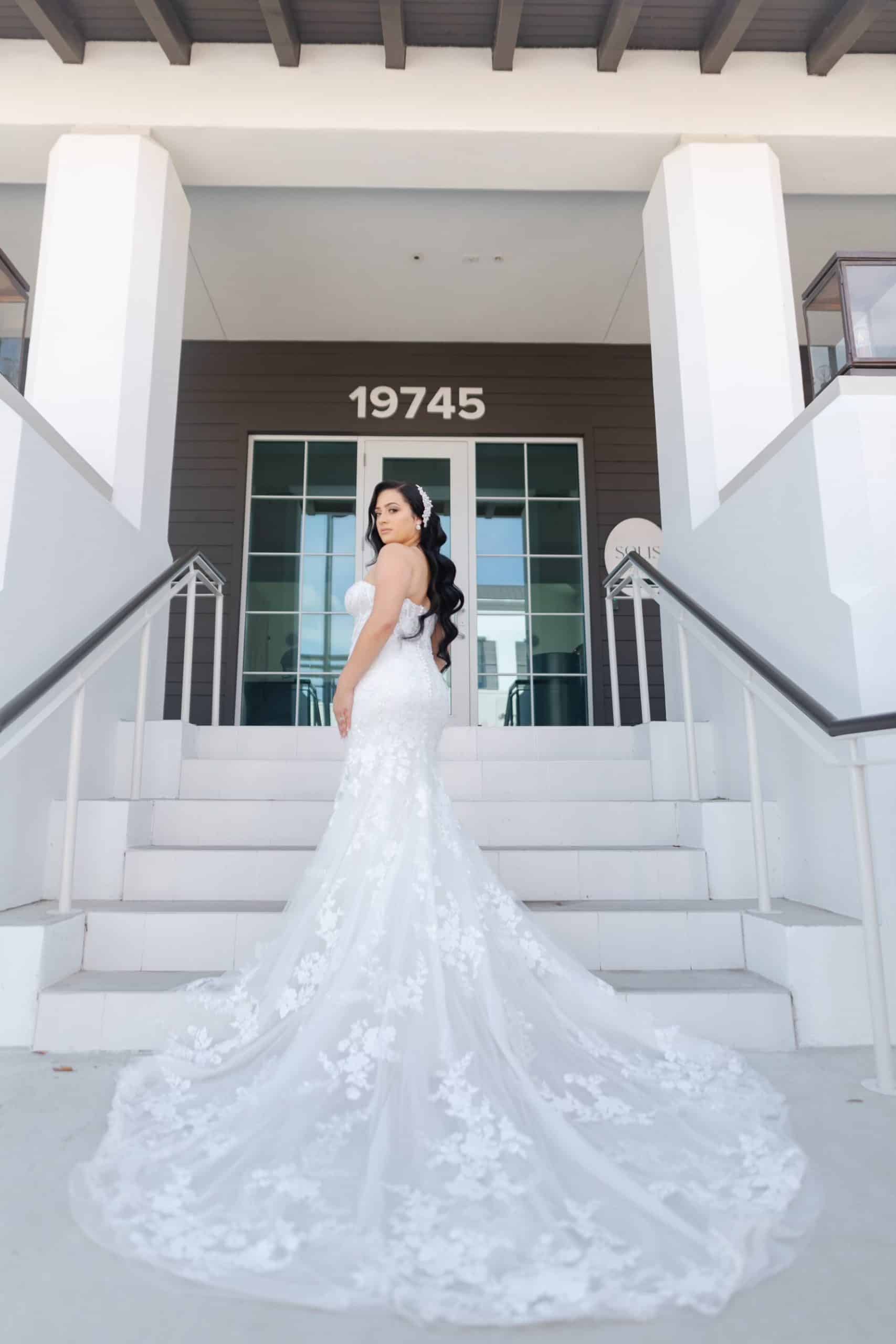 Bride posing on venue steps in a strapless lace mermaid wedding gown with a long floral train, standing beneath the building address 19745.
