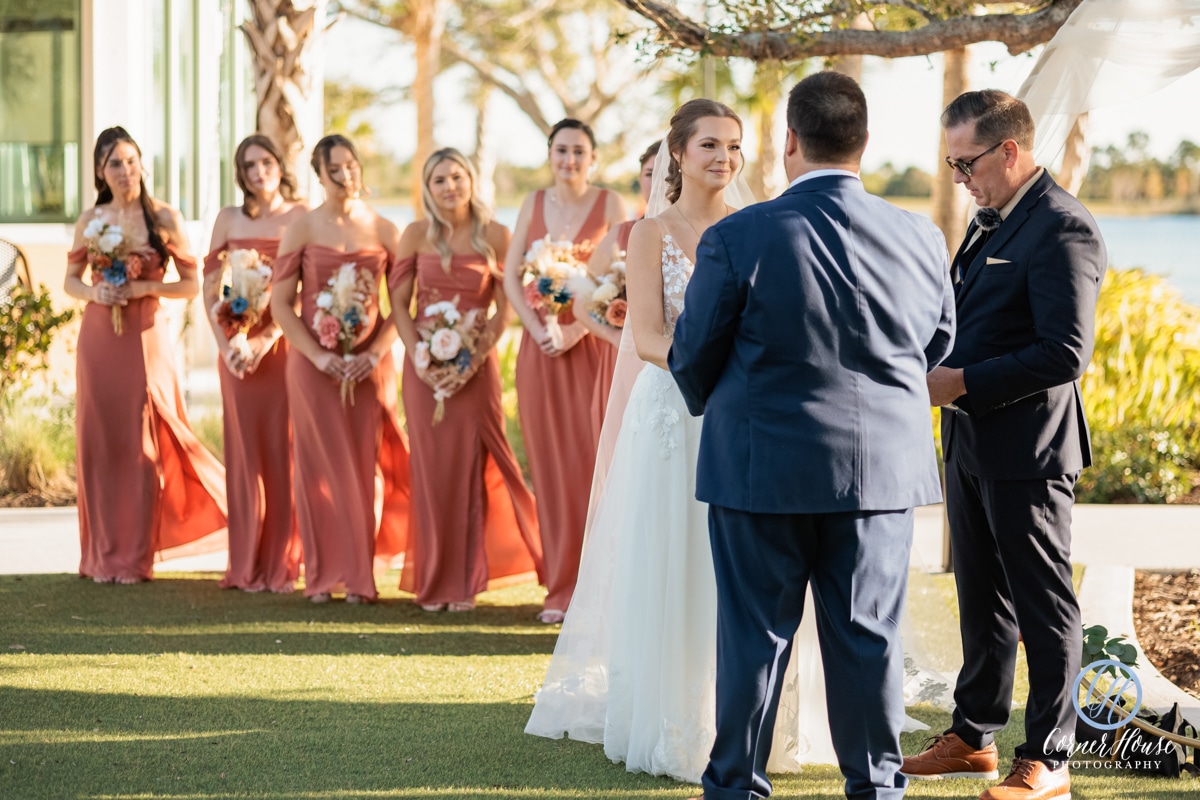 Outdoor wedding ceremony with the bride and groom exchanging vows, surrounded by bridesmaids in terracotta dresses holding floral bouquets