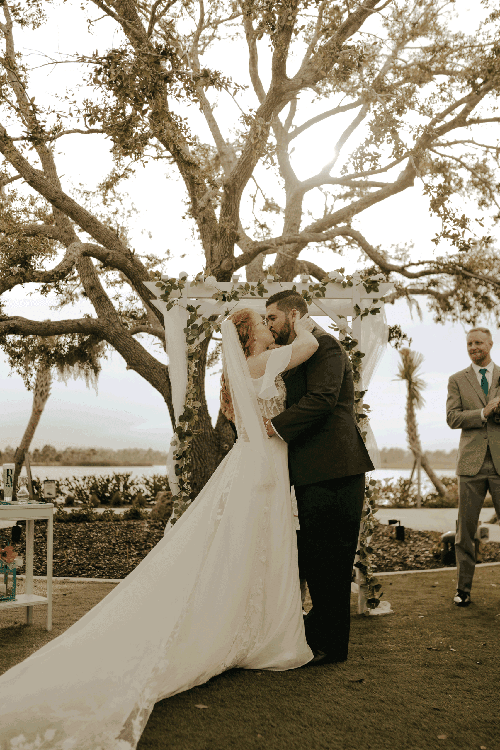 Bride and groom sharing their first kiss under a white floral-draped wedding arch at an outdoor lakeside ceremony beneath a large oak tree