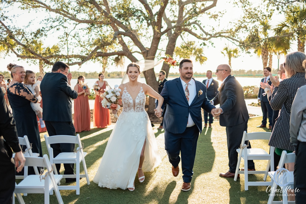 Bride and groom walking back up the aisle hand in hand during an outdoor wedding ceremony, surrounded by cheering guests and bridesmaids in terracotta dresses under a large oak tree