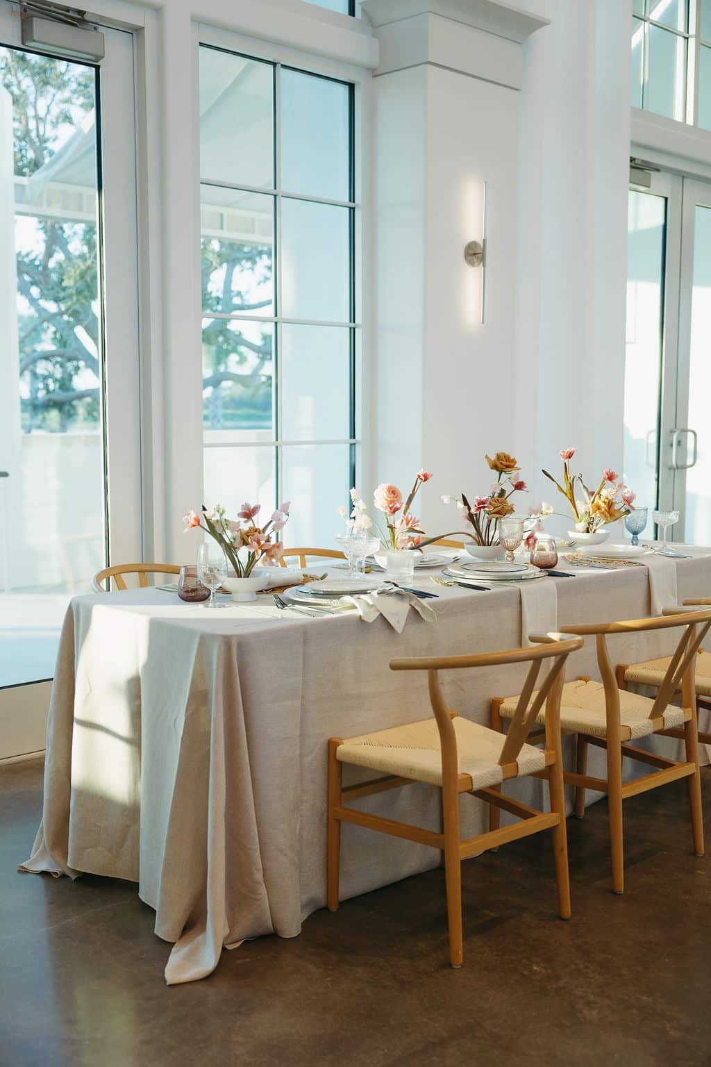 Minimalist wedding tablescape with neutral linens, modern wooden chairs, and pastel floral arrangements set inside a bright venue with large windows.