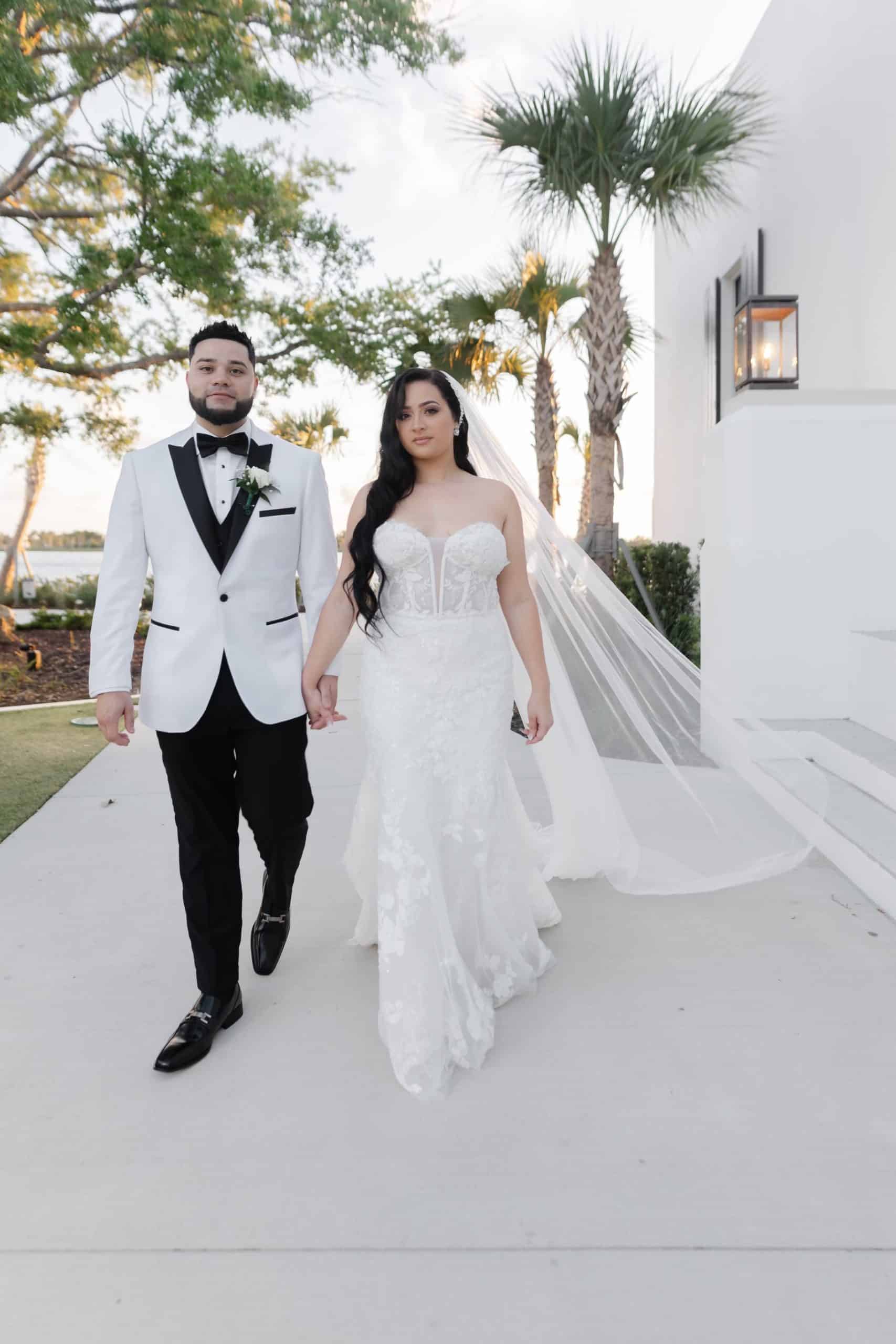 Elegant bride and groom walking hand in hand outdoors at a modern wedding venue with palm trees, the bride in a lace mermaid gown and the groom in a white tuxedo jacket.