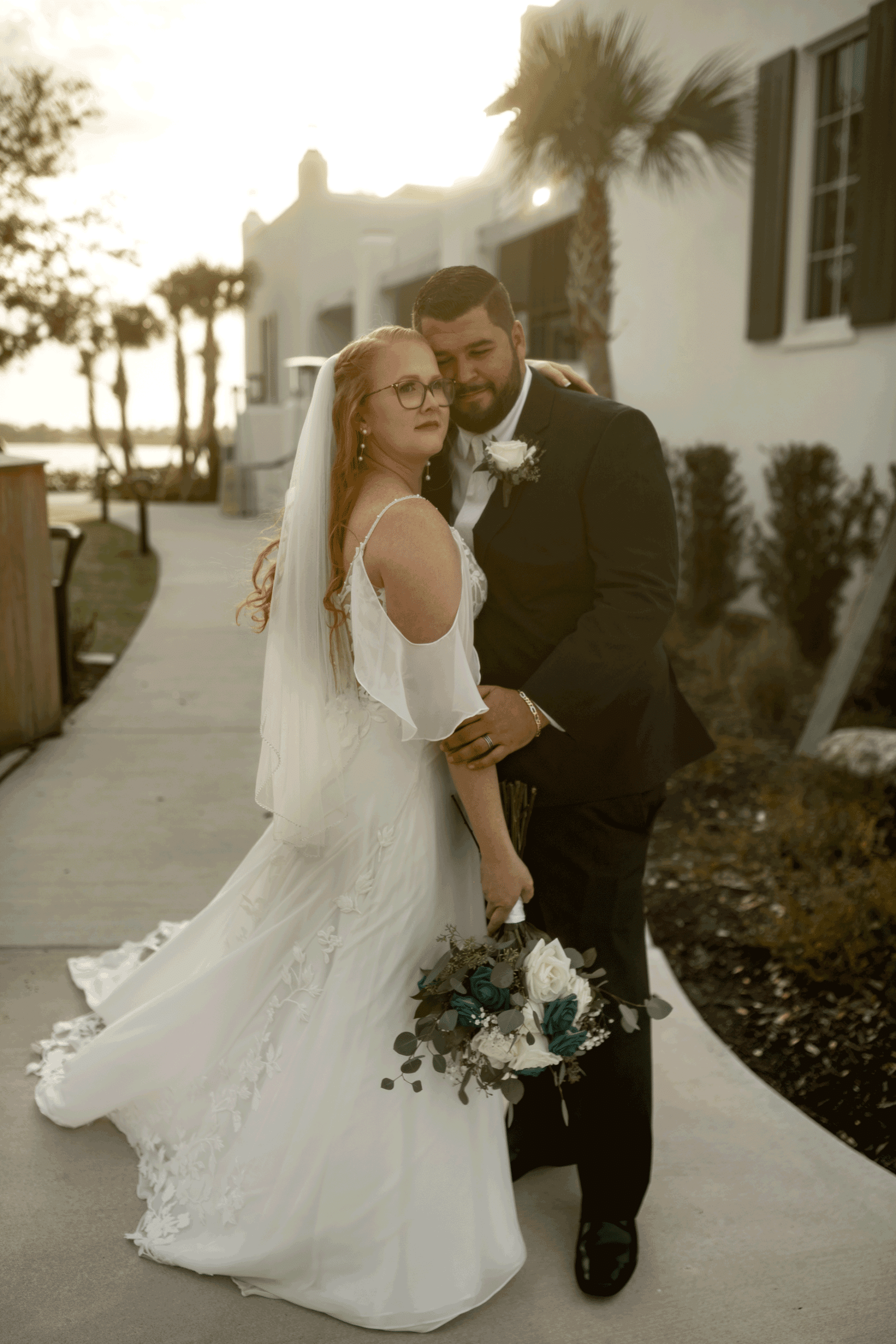 Bride and groom embracing during golden hour at an outdoor wedding venue, the bride holding a white and teal floral bouquet and wearing an off-shoulder lace gown.