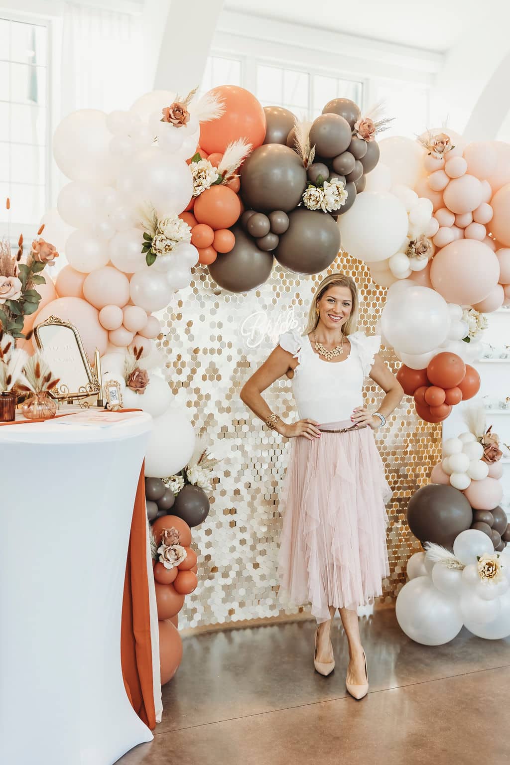 Woman posing in front of a peach, white, and brown balloon arch with floral details and a gold sequin backdrop at a decorated venice venue at Wellen Park