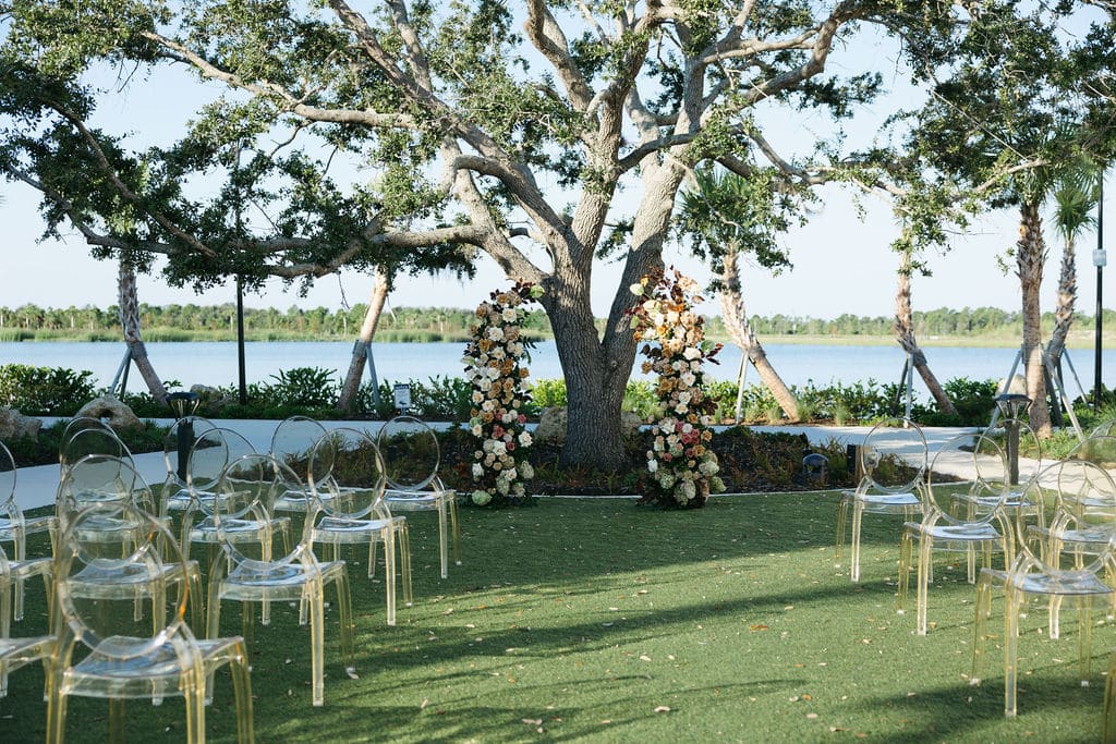 Outdoor wedding ceremony setup under a large oak tree with a floral arch and clear ghost chairs overlooking a scenic lakeside backdrop.
