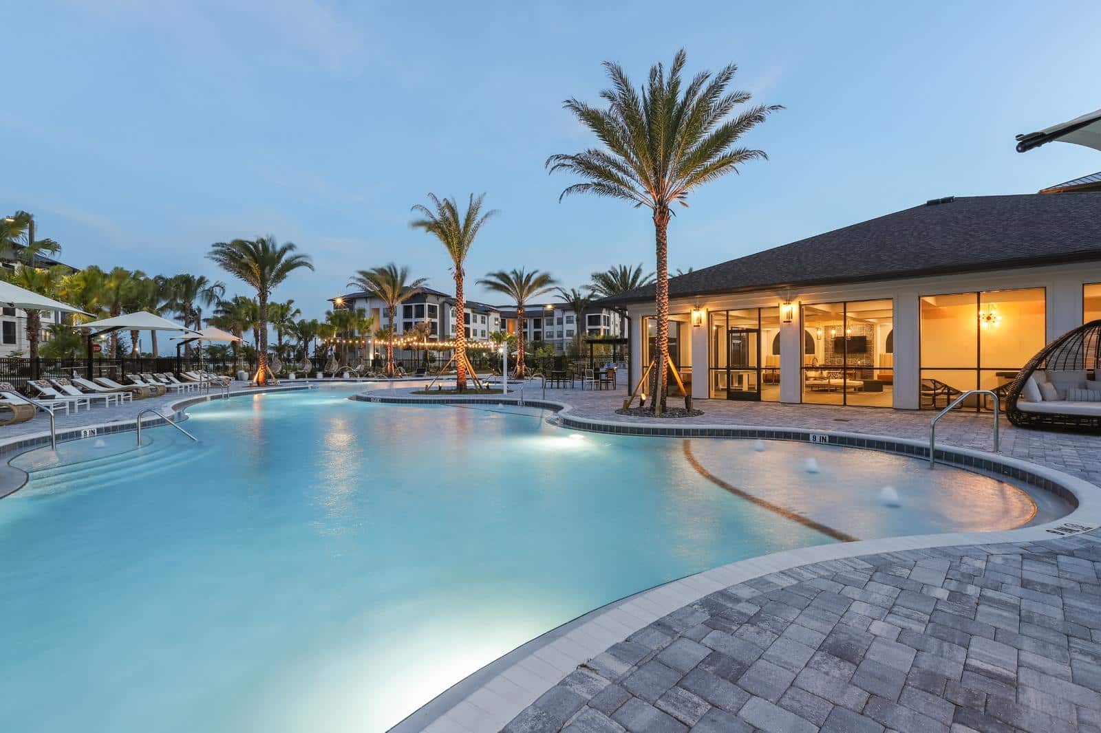 Resort-style apartment pool surrounded by palm trees, lounge chairs, and shaded umbrellas.